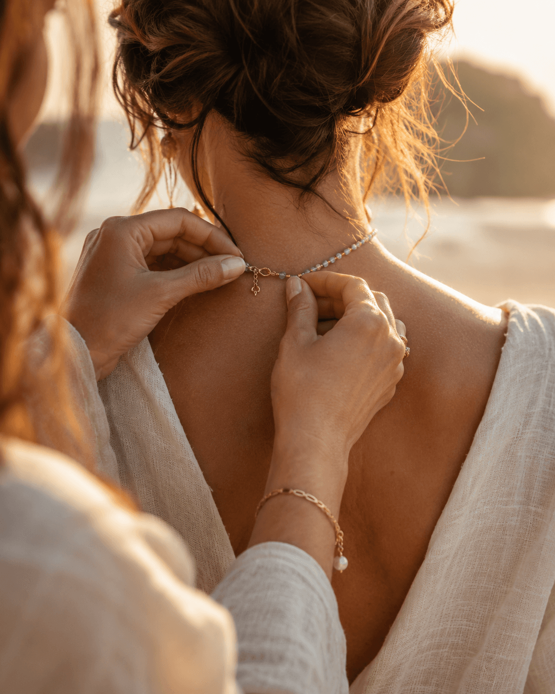 Manos abrochando un collar de aguamarina en la nuca de una mujer vestida de lino junto al mar al atardecer