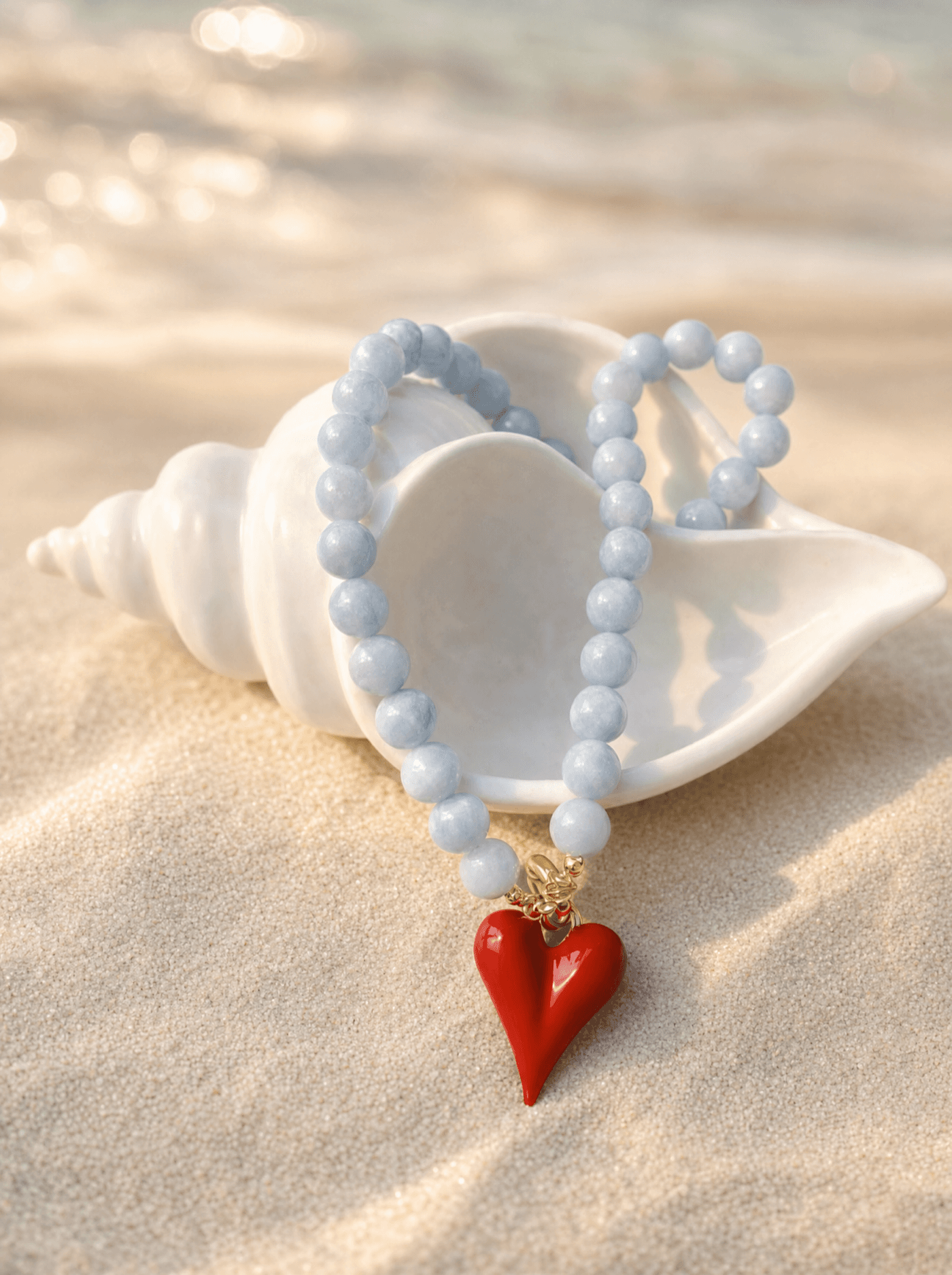 Pulsera de cuentas celestes de aguamarina con dije de corazón rojo sobre una caracola blanca.