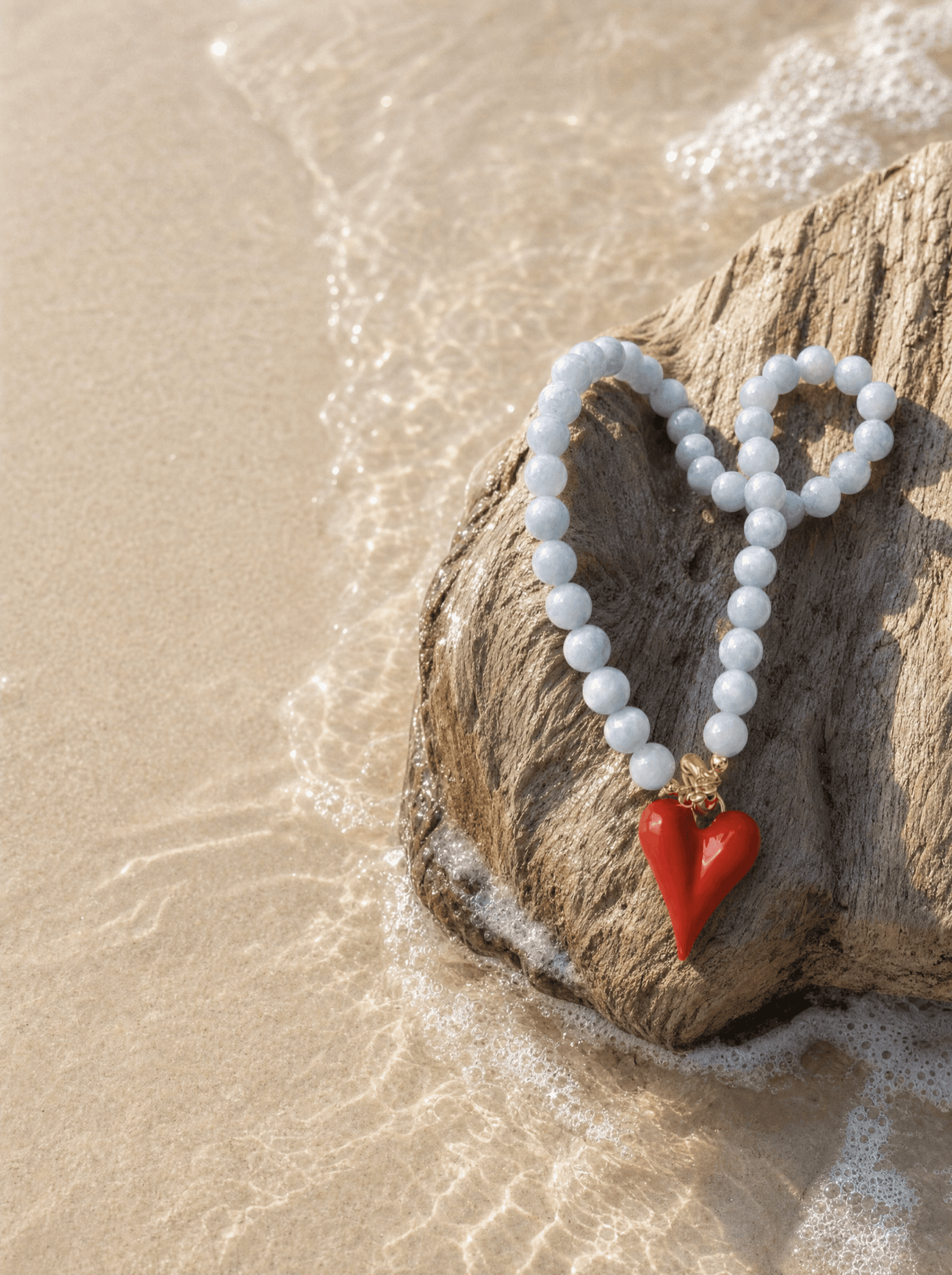 Pulsera de aguamarina con corazón rojo sobre madera de deriva a la orilla del mar al atardecer.